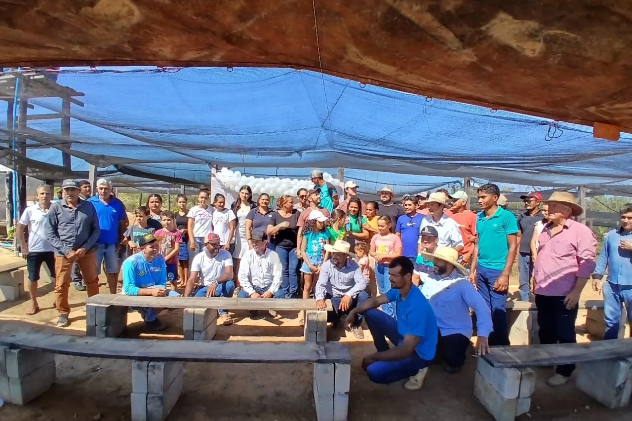 A large group of members and families posing under the nursery shade cloth, seated on wooden benches and standing.
