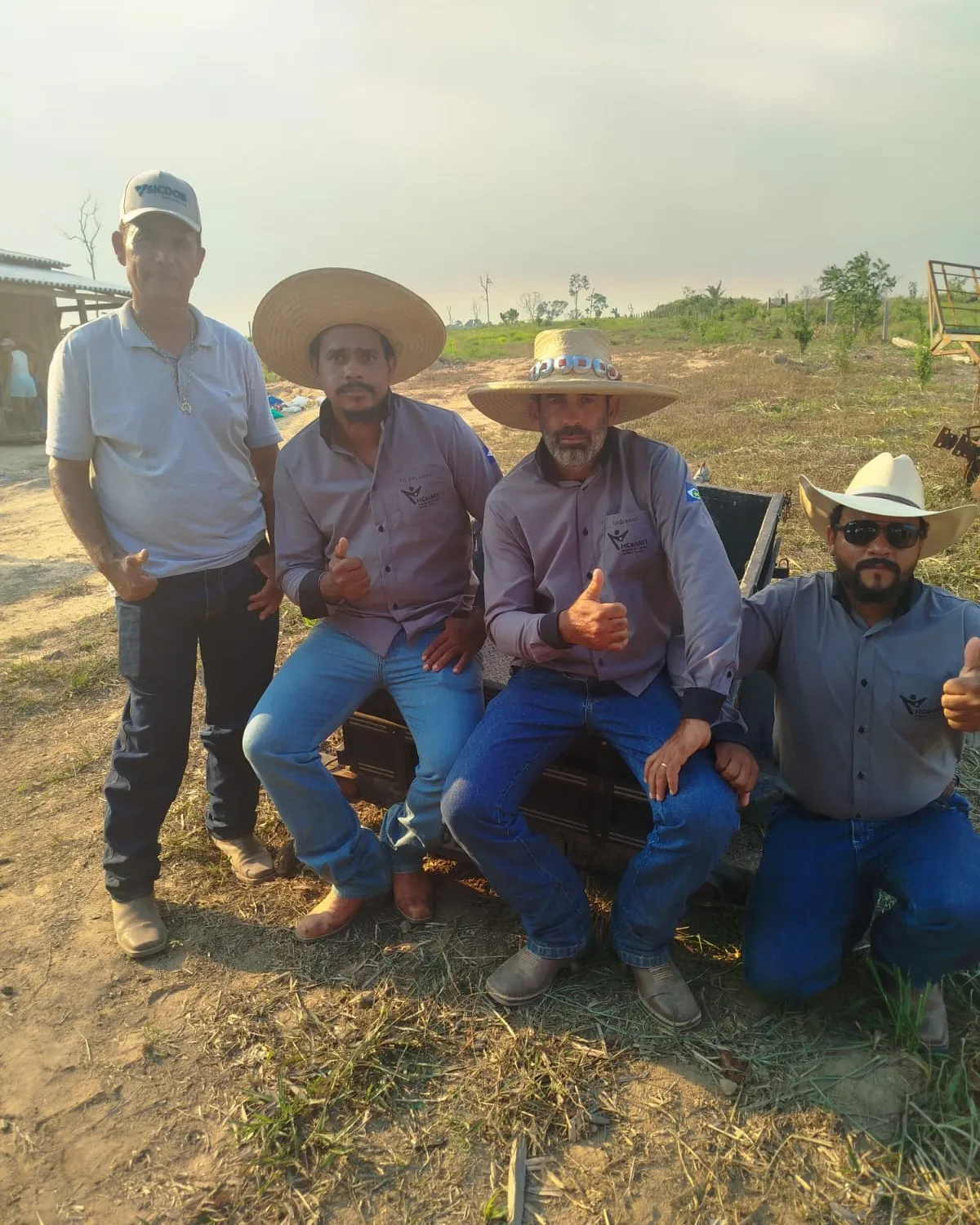 Four men in hats and button-up shirts, seated and standing beside a piece of farm equipment in an open field.