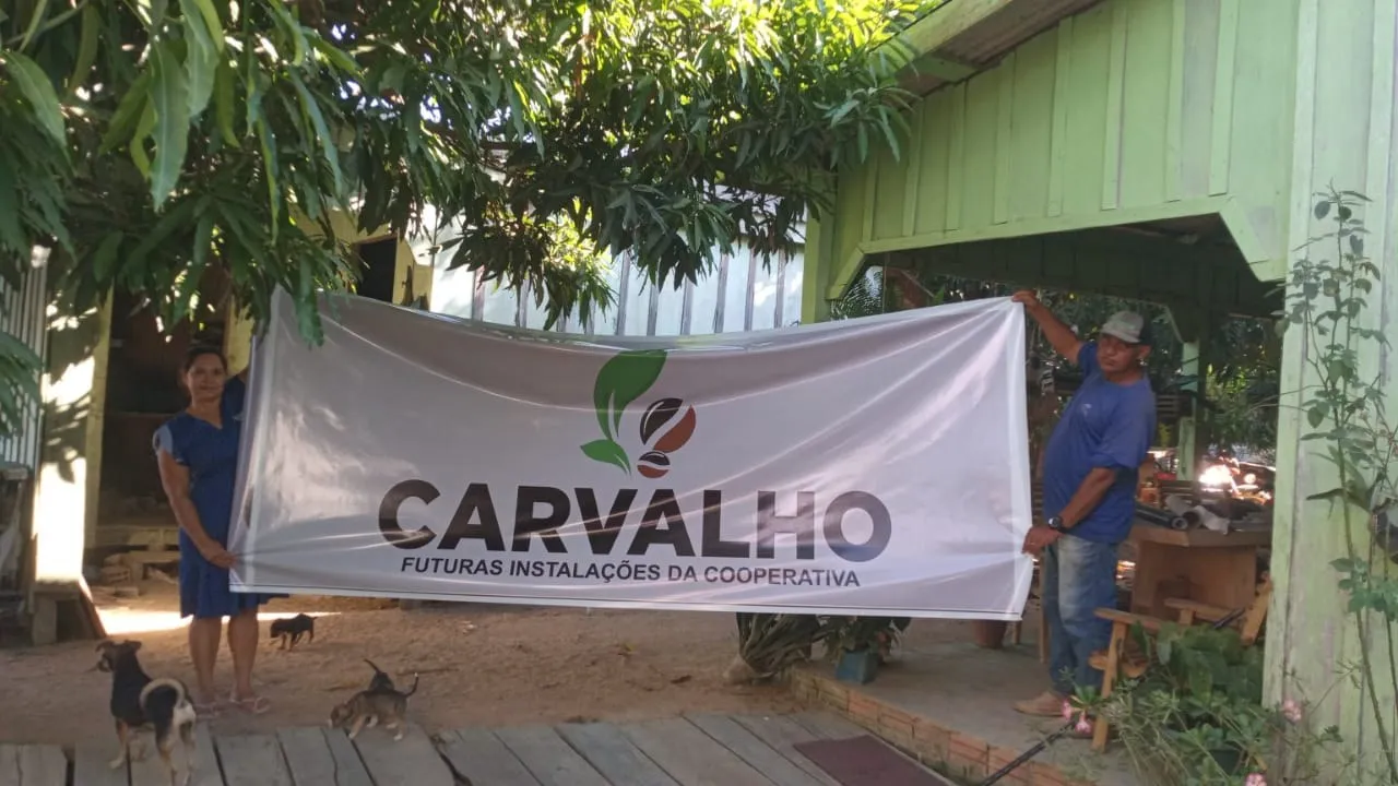 Duas pessoas segurando uma faixa branca com o logotipo Carvalho e o texto “Futuras instalações da cooperativa” em frente a uma casa de madeira.